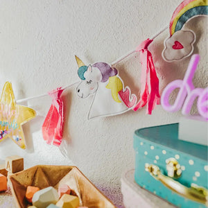 Rainbows & Unicorns Birthday Banner hanging on wall with blue suitcase and bin of wooden blocks in frontTiny Owls Gift Co