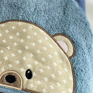 Close-up of a blue towel with a teddy bear face patch featuring a pattern of white plus signs on a beige background.
