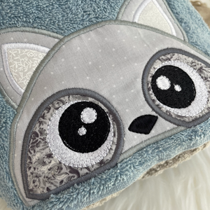 Close-up of a raccoon face appliqued with large, cartoonish eyes on a soft blue towel hood