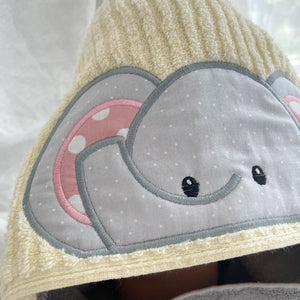 Close-up of a  yellow towel hood featuring a large applique elephant face on the front. The elephant face is made from light gray fabric with tiny white polka dots, outlined in gray, with a pink inner ear decorated with white polka dots and embroidered black eyes.