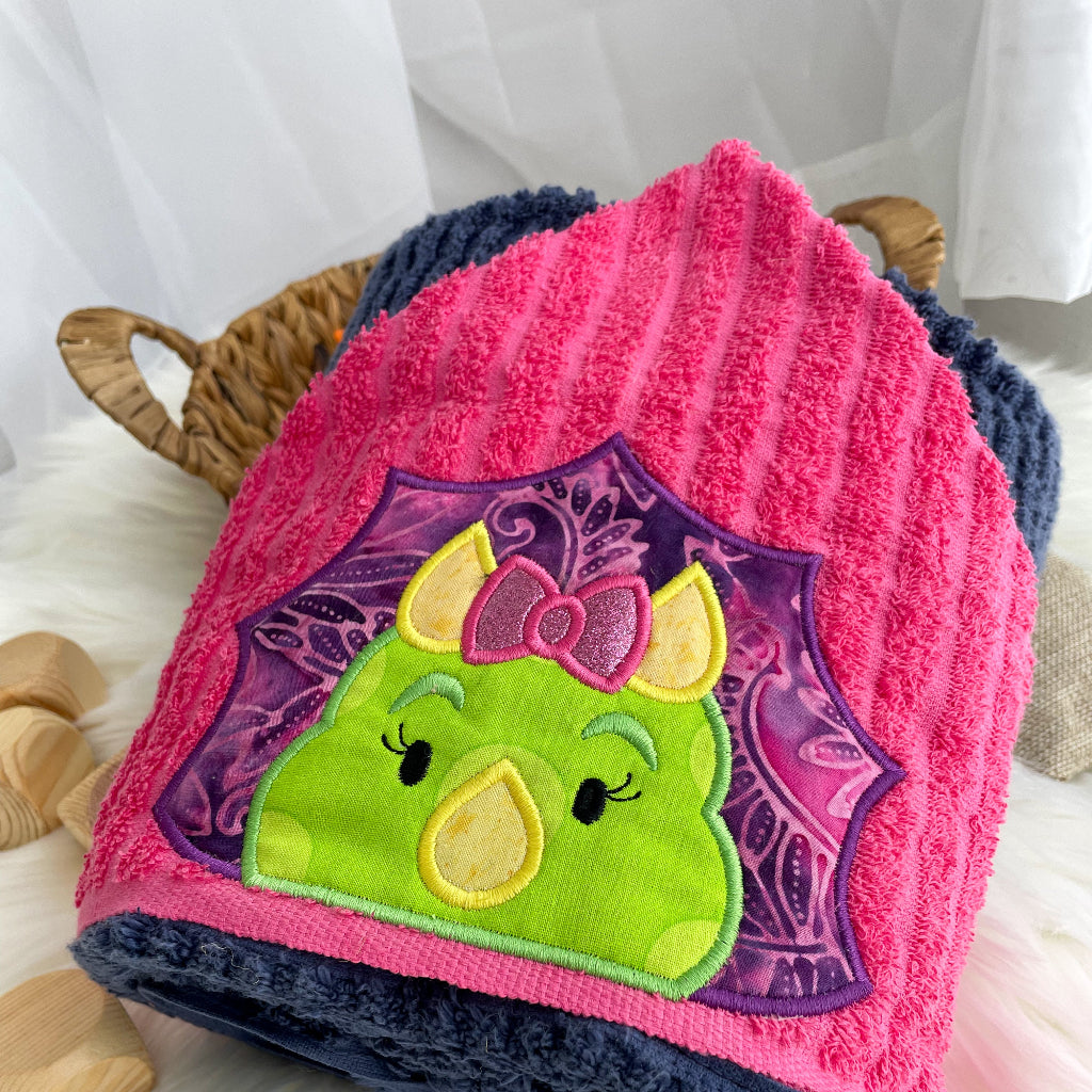 Pink and blue towel with green and purple dinosaur character and purple bow on a white background