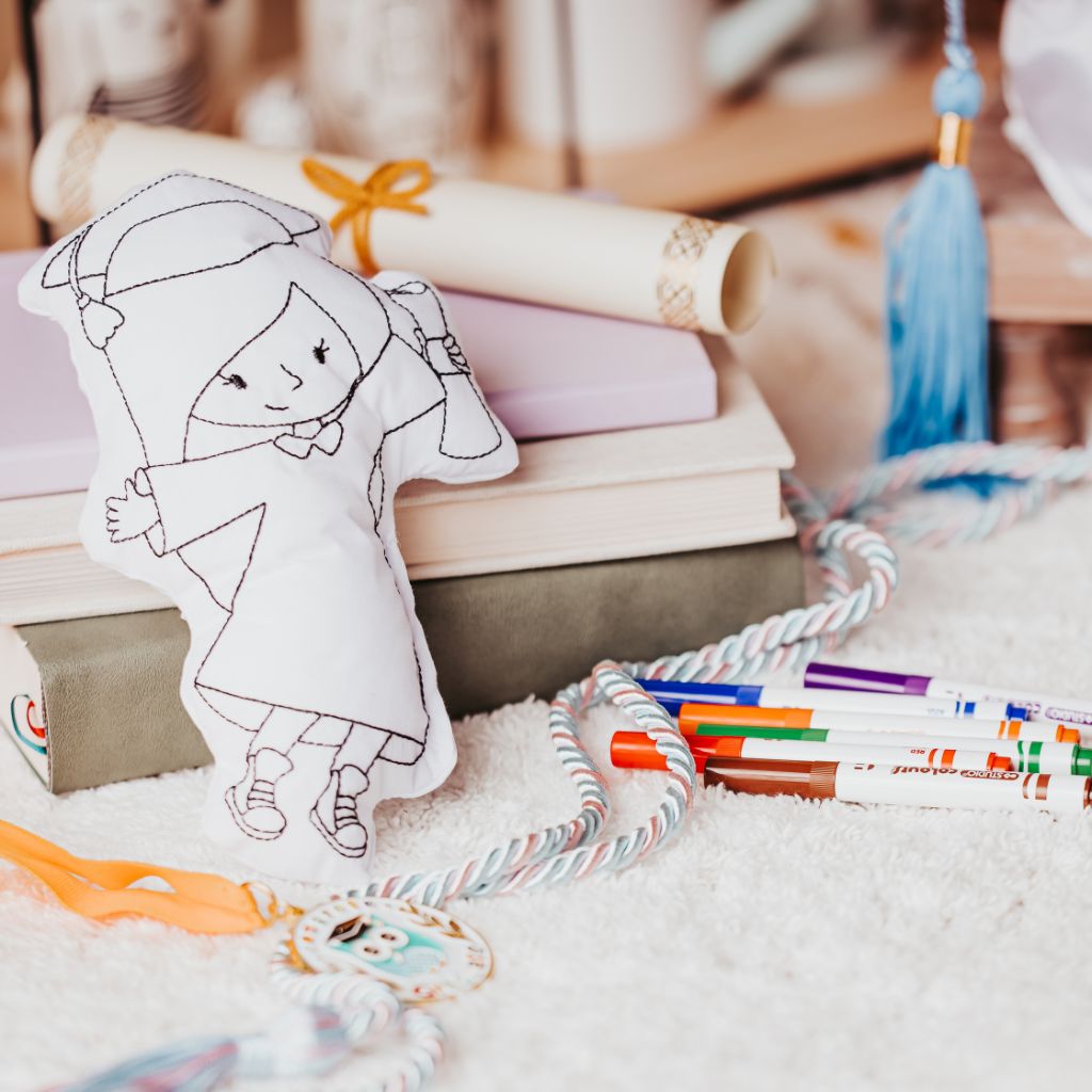 colouring pillow in the shape of a little girl holding a diploma surrounded by markers and leaning on a stack of books