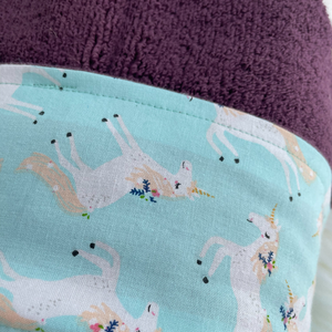Close up of a purple towel with a fabric trim featuring white unicorns on a pale blue background