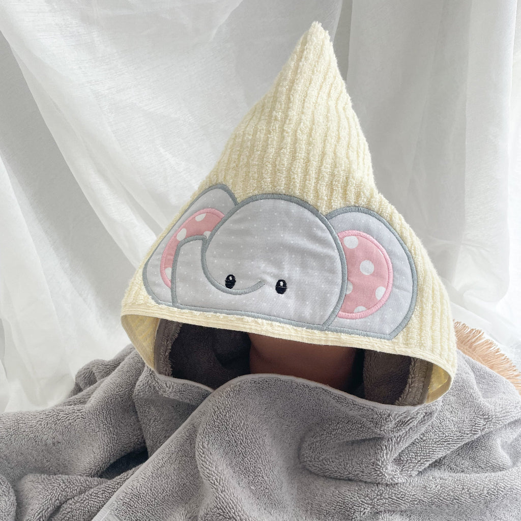 A baby wrapped in a soft gray towel, wearing a yellow towel hood with an embroidered elephant face featuring pink polka-dot ears.