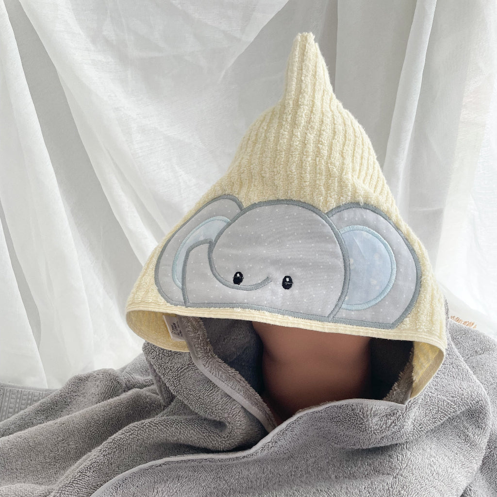 A baby wearing a pale yellow and grey  hooded towel with a gray elephant face applique on the front, set against a soft white curtain background.