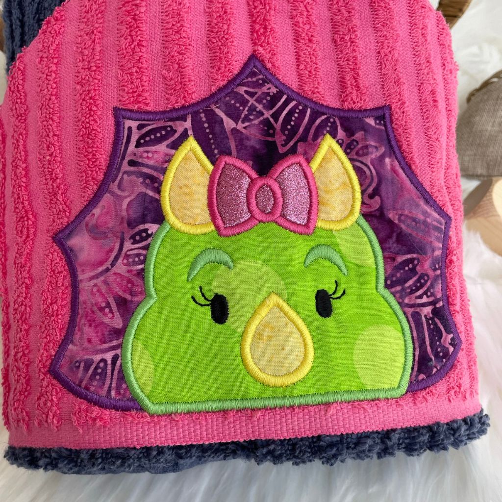 Pink and blue towel with a green and purple dinosaur patch featuring a bow on a white surface