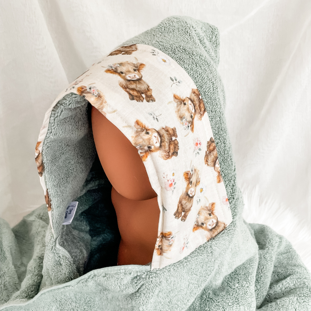 A hooded baby towel or wrap in sage green terry cloth with a printed hood panel featuring watercolor-style brown highland cows and small floral details, displayed on a mannequin head against a soft white fabric backdrop.