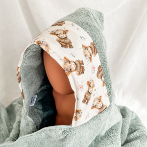 A hooded baby towel or wrap in sage green terry cloth with a printed hood panel featuring watercolor-style brown highland cows and small floral details, displayed on a mannequin head against a soft white fabric backdrop.