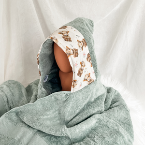A hooded baby towel or wrap in sage green terry cloth with a printed hood panel featuring watercolor-style brown highland cows and small floral details, displayed on a mannequin head against a soft white fabric backdrop.