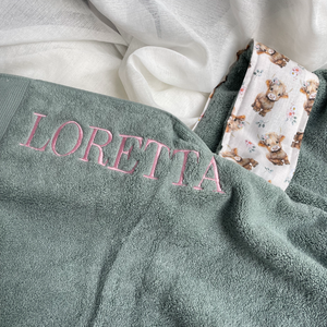 A close-up of a personalized towel in sage green terry cloth with the name 'LORETTA' embroidered in pink on the chest. fabric features a cute pattern of small brown highlnad cows with tiny colorful flowers on a cream background.
