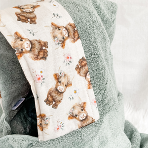 A close-up of a towel in sage green terry cloth fabric features a cute pattern of small brown highlnad cows with tiny colorful flowers on a cream background.